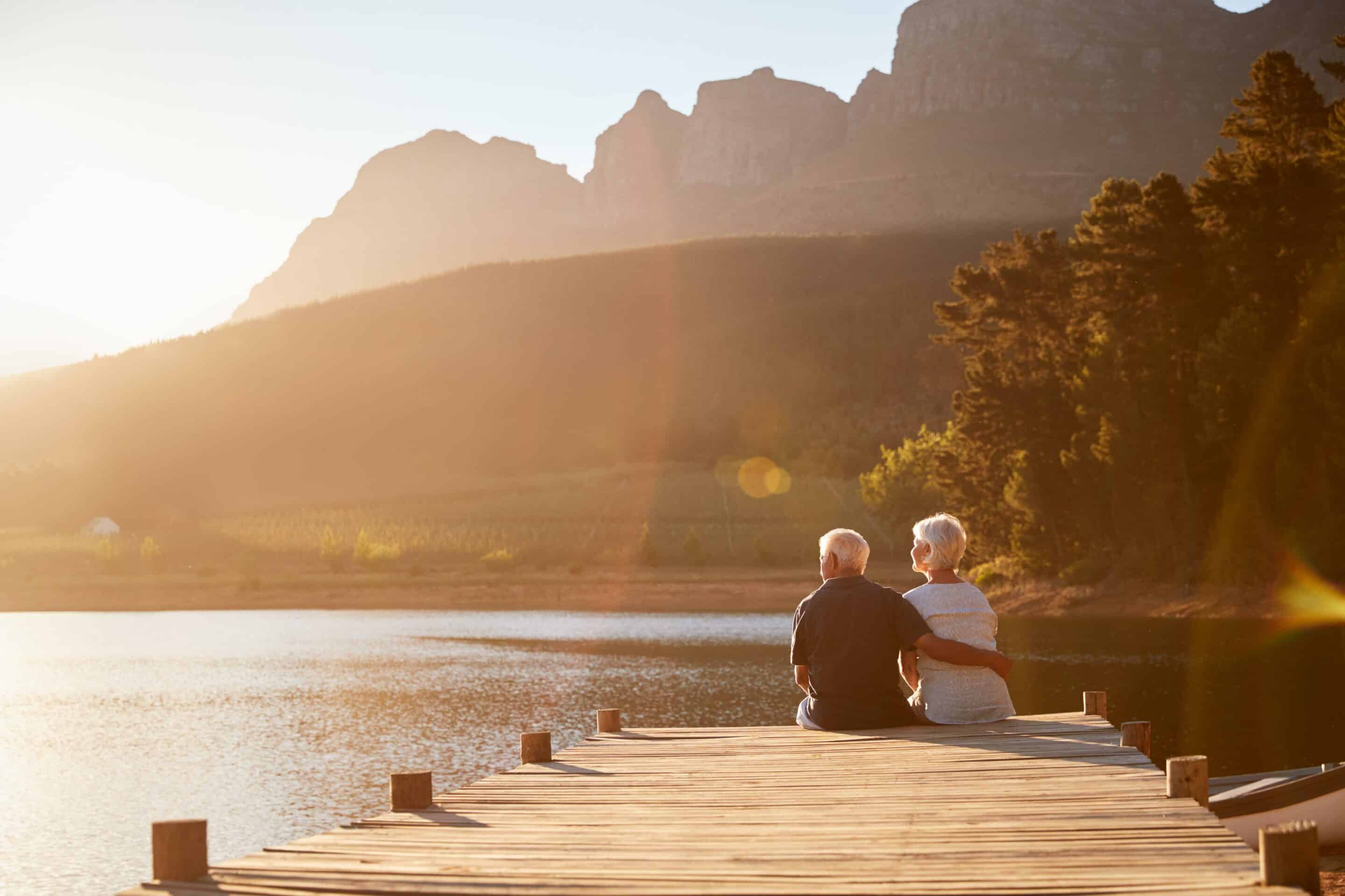 Romantic Senior Couple Sitting On Wooden Jetty By Lake And Enjoying Their Retirement Thanks To Outlook Wealth Advisors